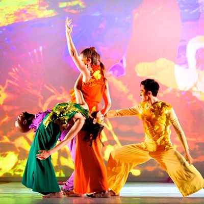 The Seldoms, arrayed in brightly colored costumes, dance in front of a screen with a kaleidoscope of colors and patterns projected on it