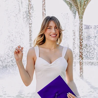 Maddie Marsman stands in front of Frog Fountain with a purple mortarboard in one hand and makes the Go Frogs sign with the other