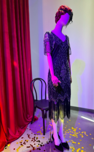 A mannequin wearing a black beaded evening gown with a formfitting silhouette. It features an off-the-shoulder neckline with a draped, asymmetrical design.