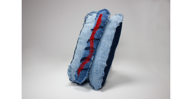 A sculptural work by Chris Wicker features folded, frayed blue denim with a prominent strip of red stitching running vertically through the center, photographed against a neutral gray background.