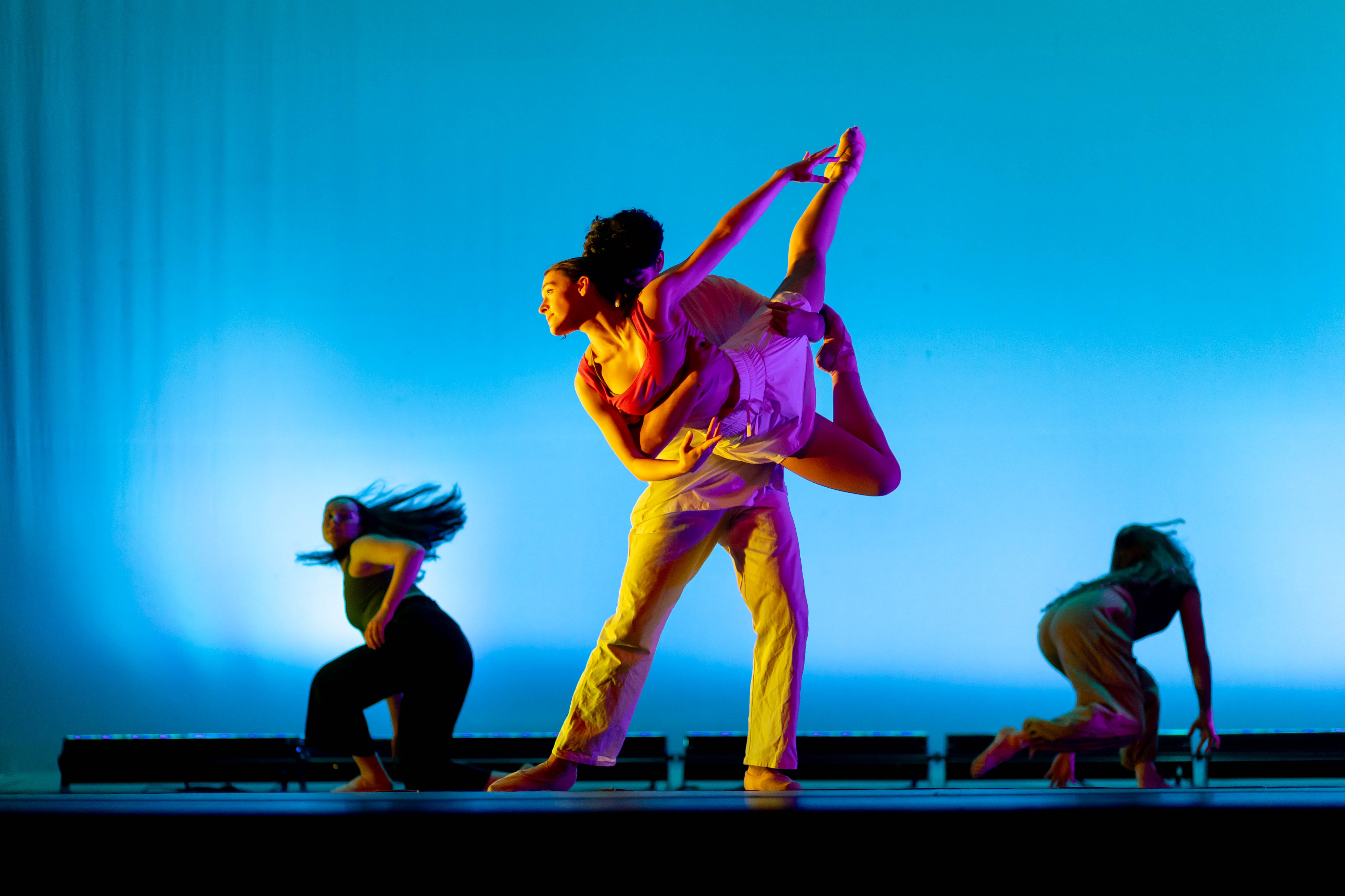 Four dancers perform onstage against a vivid blue backdrop illuminated with purple and amber lighting. At center, one dancer lifts and supports another in an extended pose while two additional dancers move on either side, creating a dynamic contemporary dance scene.
