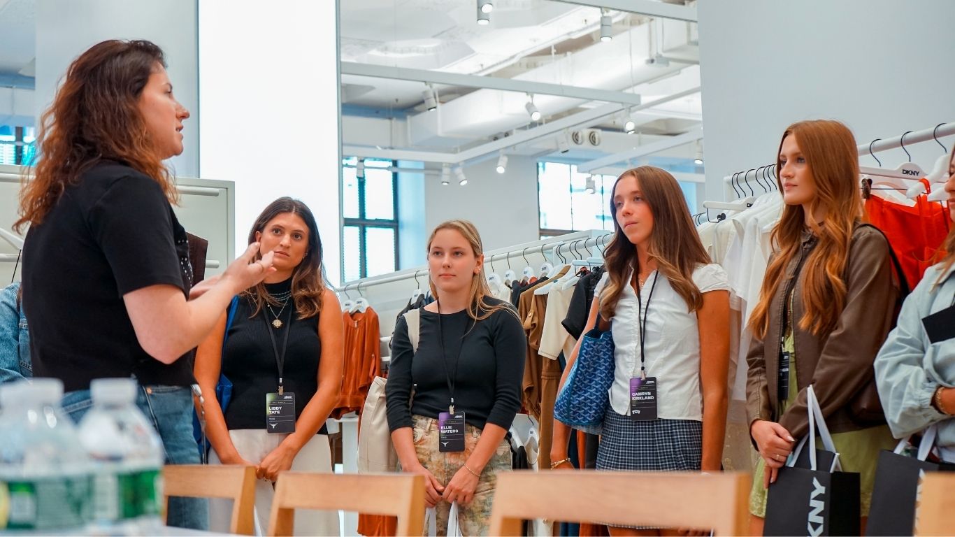Camryn Kirkland, wearing a light blue shirt and plaid skirt, stands with other CLASS participants as a fashion industry professional speaks to the group in a showroom surrounded by clothing racks.