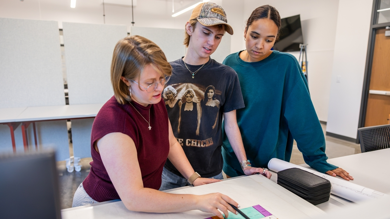Three TCU design students gather around a large worktable, examining plans and color samples while discussing a project.
