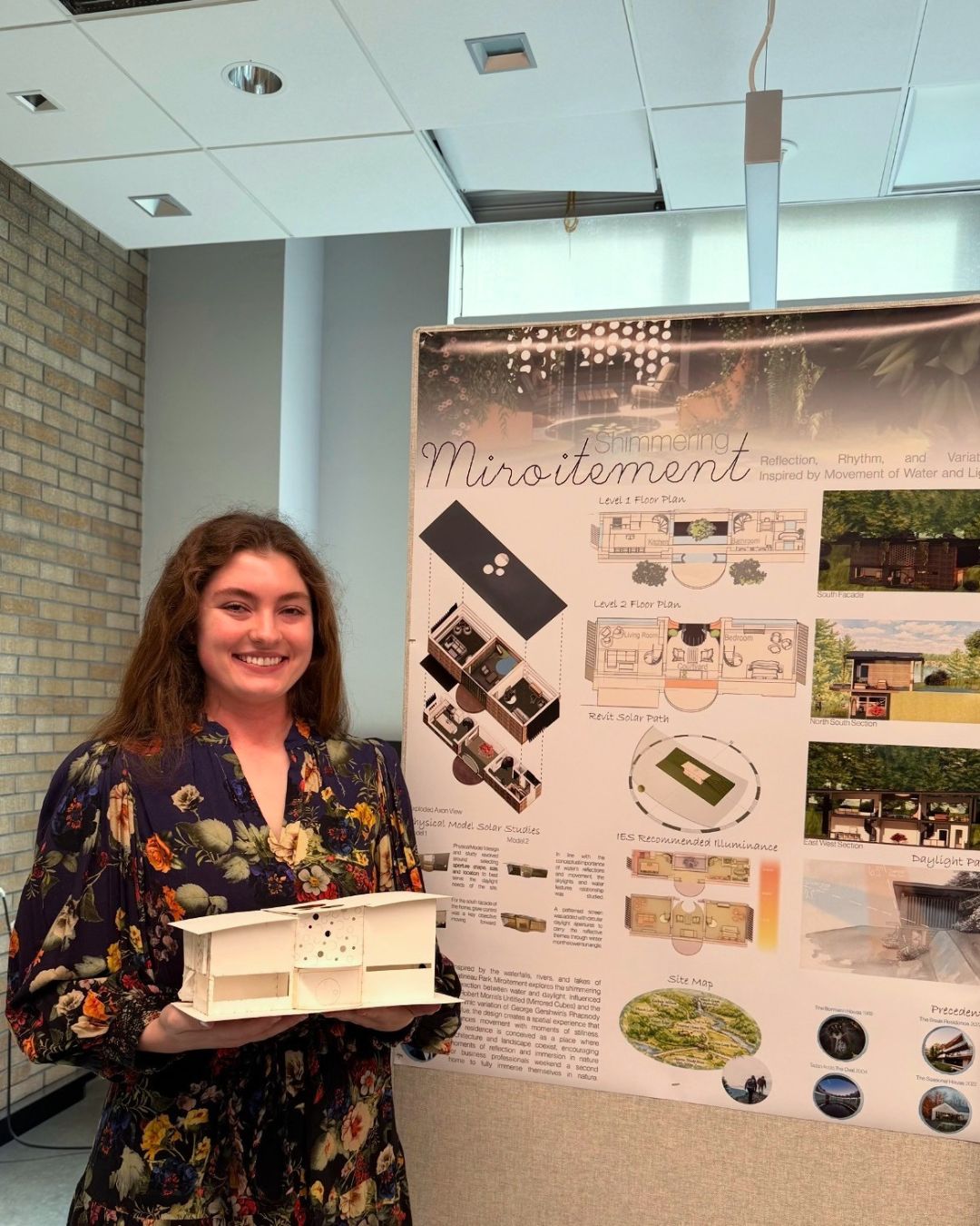Lily Plaster smiles while holding a white architectural model beside a presentation board displaying floor plans, renderings, lighting studies and sustainability considerations for her design project.
