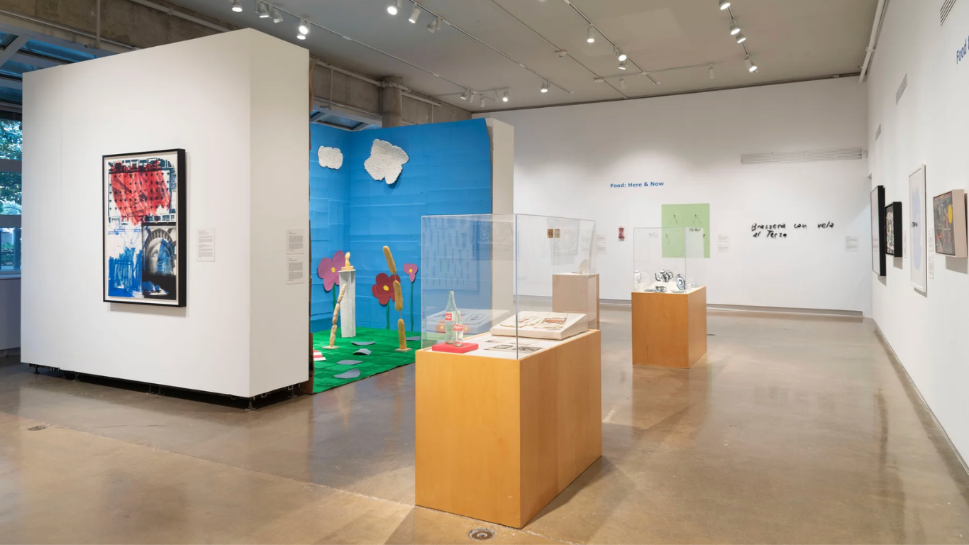 A wide view of What’s for Dinner? at TCU’s Moudy Gallery shows framed artwork, display cases and sculptural installations arranged throughout the gallery, including a colorful immersive installation with a blue backdrop and oversized flowers and food forms.