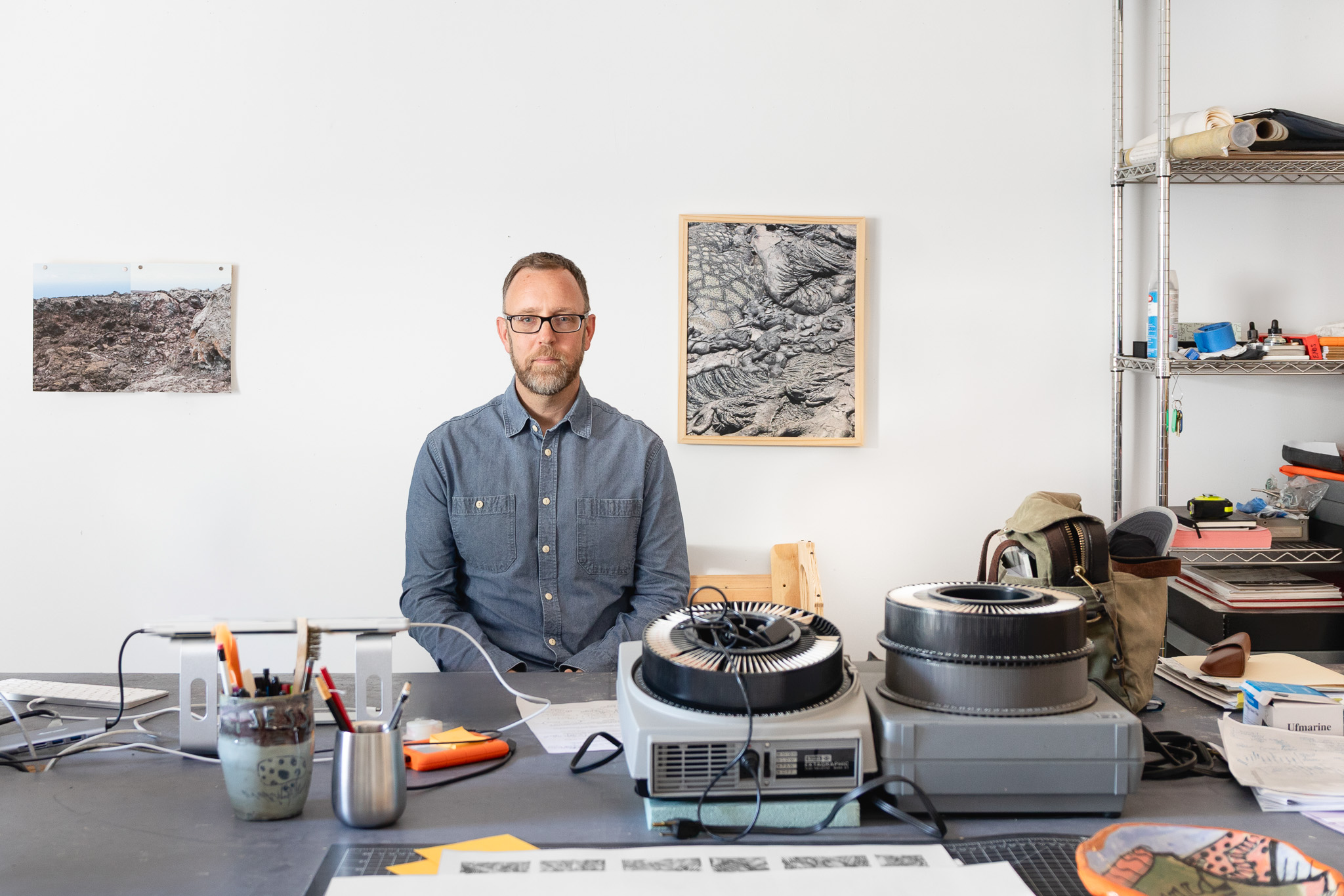 James Enos sits behind a worktable in his art studio, surrounded by drawing tools, equipment and supplies. Photographs and a framed black-and-white artwork hang on the wall behind him.
