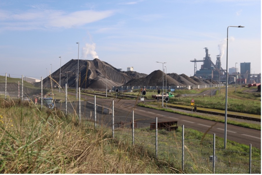 Large mounds of dark industrial material rise beside roads and railway tracks at Tata Steel in the Netherlands, with factory structures and smokestacks visible in the background.