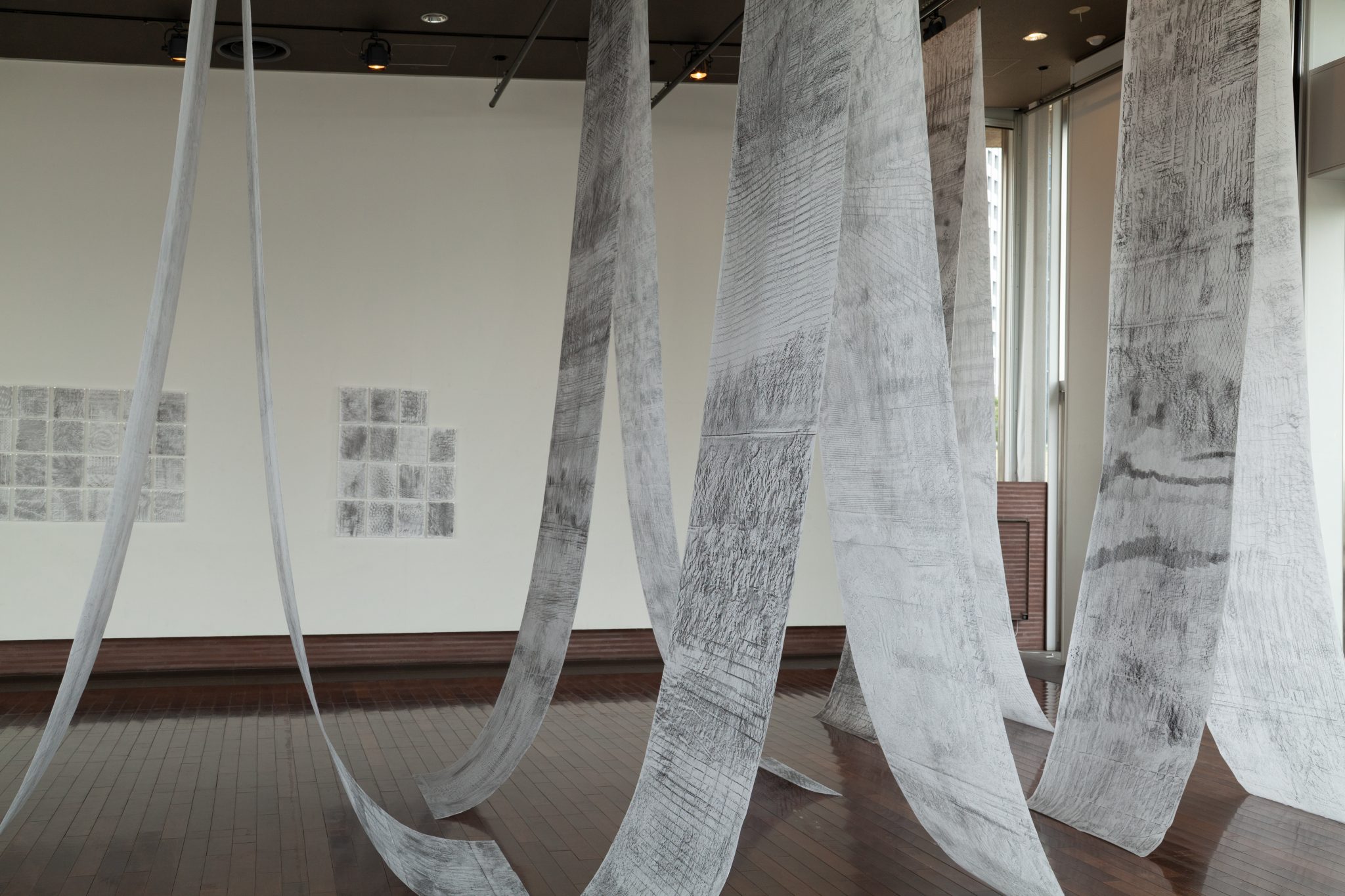 Long, suspended sheets of white material covered in intricate gray markings hang from the ceiling of a gallery, forming sweeping curves that extend toward the wooden floor. Additional drawings are displayed on the surrounding walls.