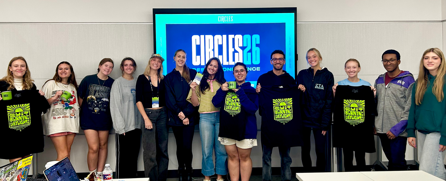 A group of TCU Design Focus students poses in front of a large Circles 2026 Conference screen in the Fine Arts Building's Jury Room. Several students hold black T-shirts with bright green graphics and conference merchandise.