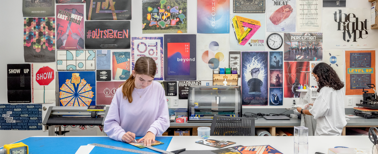 Two TCU graphic design students work in a studio filled with colorful posters and graphic design projects. One student uses a ruler and cutting tools at a large worktable while another works with equipment in the background.