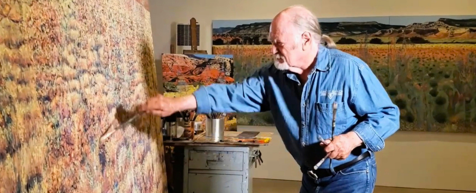 Jim Woodson, dressed in a denim work shirt and jeans, paints a large landscape canvas covered in dense, sun-lit grasses. He reaches across the surface with a wide brush while holding several other brushes in his left hand. A metal rolling cart of paints and brushes and additional finished landscapes fill the softly lit studio behind him.