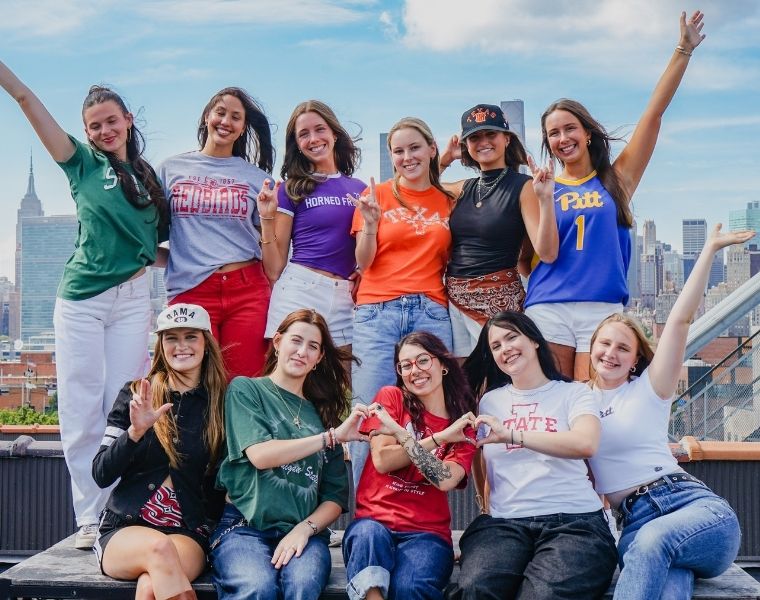 Ten CLASS: The NYC Experience participants representing different universities pose together on a rooftop overlooking the Manhattan skyline. Kirkland stands in the back row wearing a purple TCU Horned Frogs shirt and making the Horned Frog hand sign.