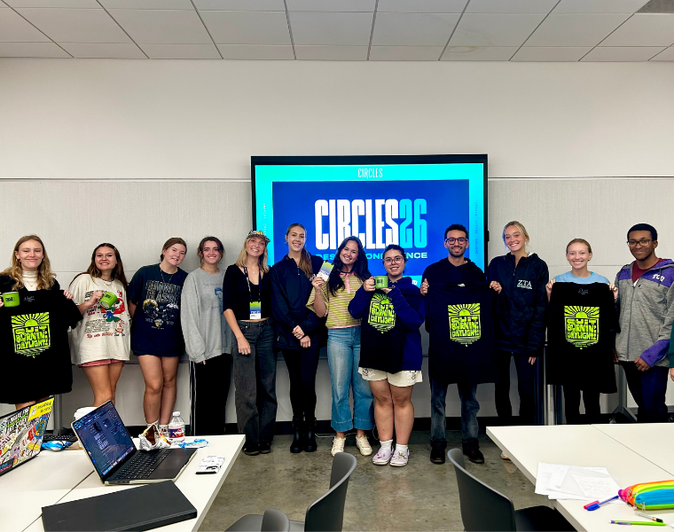 A group of TCU Design Focus students poses in front of a large Circles 2026 Conference screen in the Fine Arts Building's Jury Room. Several students hold black T-shirts with bright green graphics and conference merchandise.