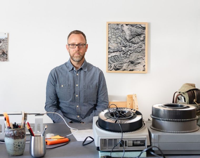 James Enos sits behind a worktable in his art studio, surrounded by drawing tools, equipment and supplies. Photographs and a framed black-and-white artwork hang on the wall behind him.