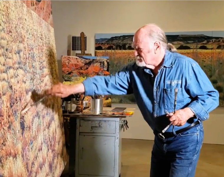 Jim Woodson, dressed in a denim work shirt and jeans, paints a large landscape canvas covered in dense, sun-lit grasses. He reaches across the surface with a wide brush while holding several other brushes in his left hand. A metal rolling cart of paints and brushes and additional finished landscapes fill the softly lit studio behind him.