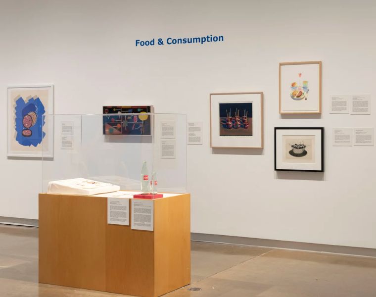 The “Food & Consumption” section of What’s for Dinner? features framed artworks and interpretive labels arranged along a white gallery wall. A wooden pedestal and glass display case in the foreground hold additional food-related objects.