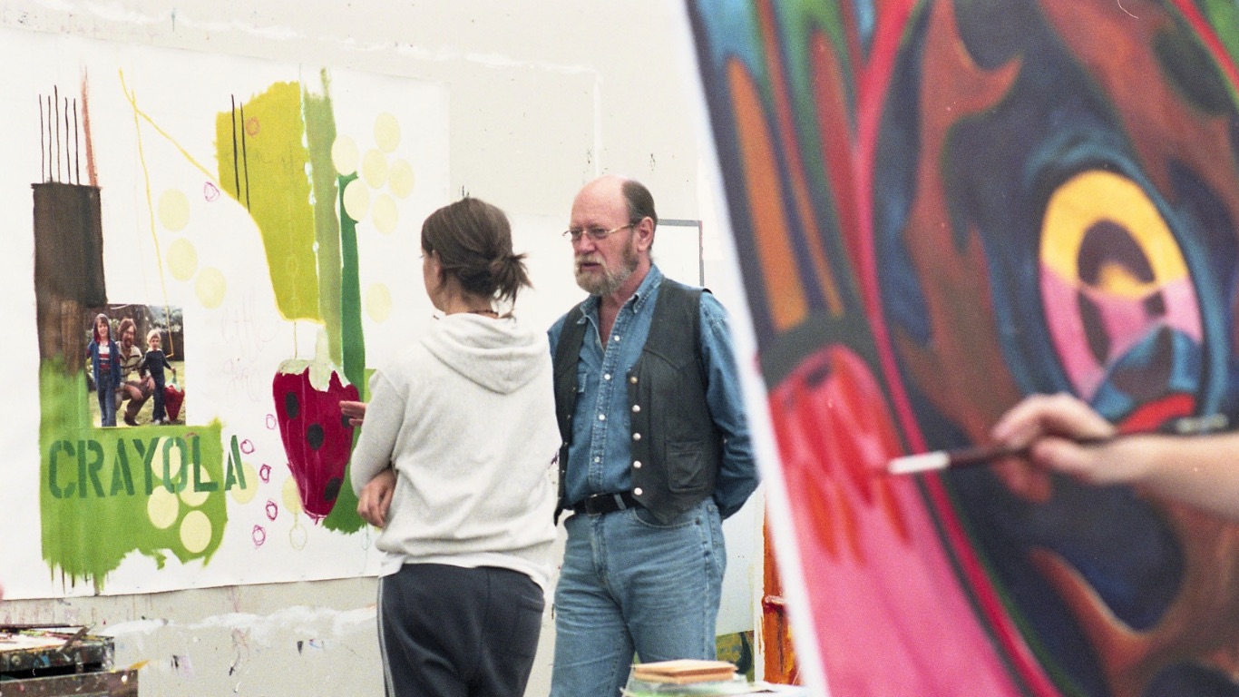 Jim Woodson in a denim shirt and a leather vest stands beside a student who is studying her mixed-media canvas. The student’s artwork features vivid green shapes, a photo collage, and the word “CRAYOLA.” In the blurred foreground, another painter’s hand works on a bold, abstract canvas, framing the scene of an informal critique.