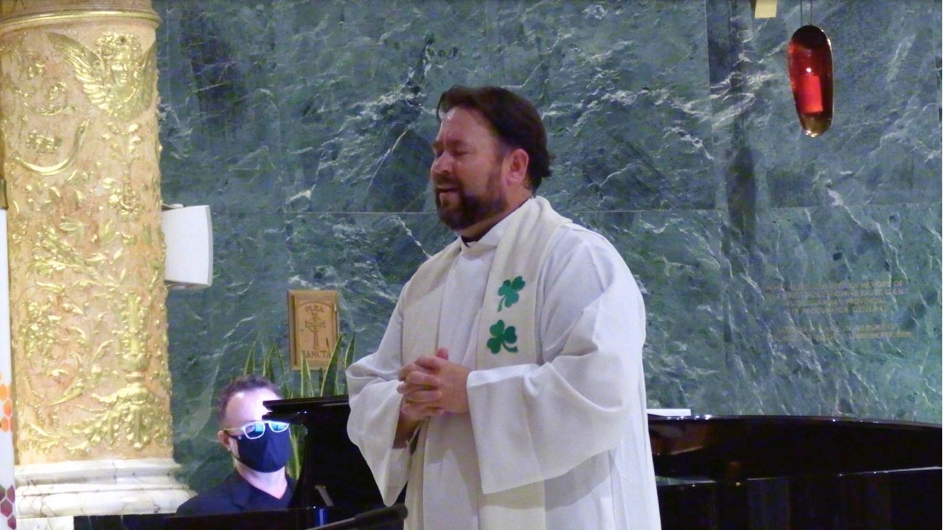 In a marble-walled sanctuary, baritone Dr. Richard Novak sings with eyes closed, wearing a white liturgical alb and stole adorned with green shamrocks. Behind him, Dr. Joey Martin sits at a grand piano, masked and partly hidden by a gilded column carved with angelic motifs.