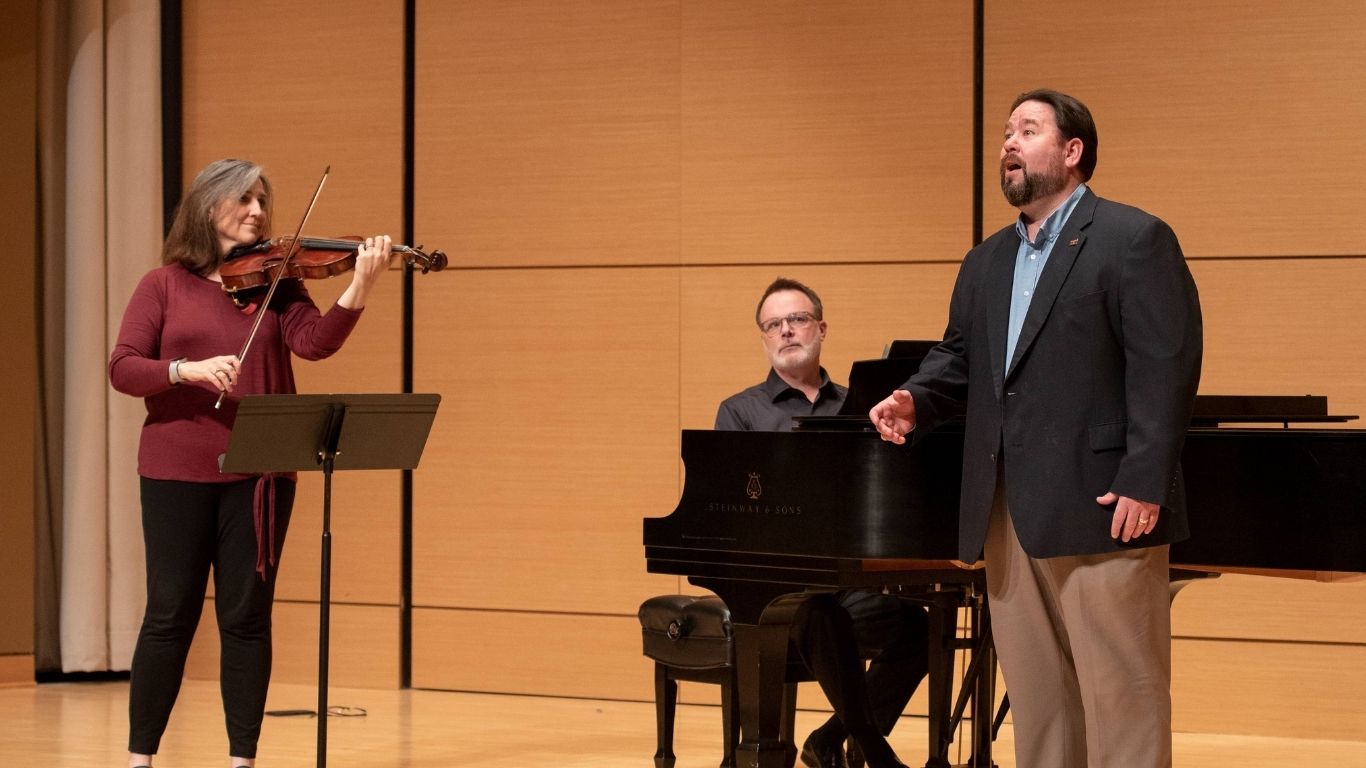 On a warm-toned recital-hall stage, violist Dr. Ames Asbell (left) plays while standing at a music stand, Dr. Joey Martin (center) accompanies on a black Steinway grand piano, and Dr. Richard Novak (right) sings in a dark blazer and open-collar shirt.