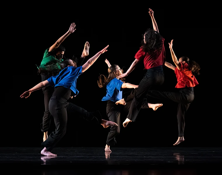Dancers wearing bright, primary colors leap in a tight group on a darkened stage