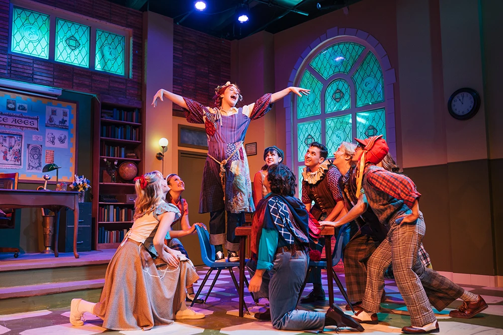 Characters encircle a female lead who is standing on a chair with outstretched arms