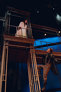 Juliet, a young woman in a long nightgown, gazes lovingly down at Romeo, a young man, climbing up iron piping to her balcony. 
