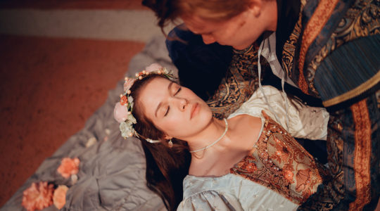 Juliet, a young woman in period dress with a floral headband, is held by Romeo, a young man, as he believes her to be dead.