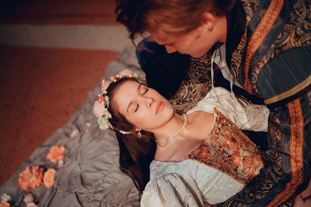 Juliet, a young woman in period dress with a floral headband, is held by Romeo, a young man, as he believes her to be dead. 