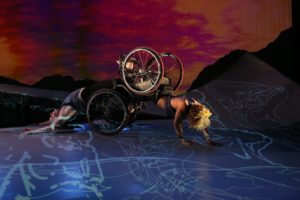 Alice, a multiracial Black woman with short curly golden hair, wearing a sleeveless tank top, is crawling on her forearms with her knees in Laurel's footplate. Laurel, a pale white woman also wearing a sleeveless top, is arching her back on the ground as she is dragged along the floor. Their wheelchair wheels are stacked and shine in the light. The sky in the background is full of deep red and orange sunset hues; blue and white projections of figures and constellations fall across the ramp and the floor. Photo by Chris Cameron, courtesy MANCC.