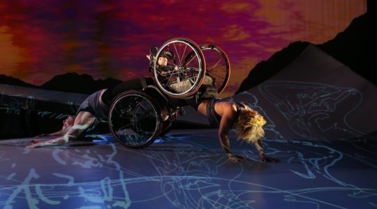 Alice, a multiracial Black woman with short curly golden hair, wearing a sleeveless tank top, is crawling on her forearms with her knees in Laurel's footplate. Laurel, a pale white woman also wearing a sleeveless top, is arching her back on the ground as she is dragged along the floor. Their wheelchair wheels are stacked and shine in the light. The sky in the background is full of deep red and orange sunset hues; blue and white projections of figures and constellations fall across the ramp and the floor. Photo by Chris Cameron, courtesy MANCC.