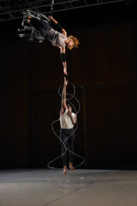 Alice Sheppard, a light-skinned multiracial Black woman with short curly hair, suspends majestically from the ceiling; one arm clasped to a cable that hooks onto her wheelchair, and another outstretched straight down towards the floor. Jerron Herman, a medium-height dark-skinned Black man with a beard and kinky high top black hair, jumps up towards Alice’s outstretched hand. He is looking directly up at her and his right and is just shy of meeting hers. His palsied left arm clings to his chest. Silver barbed wire cascades all around Jerron’s body, encircling him in a sculptural framework. Photo by Grace Kathryn Landefeld, courtesy Jacob’s Pillow.