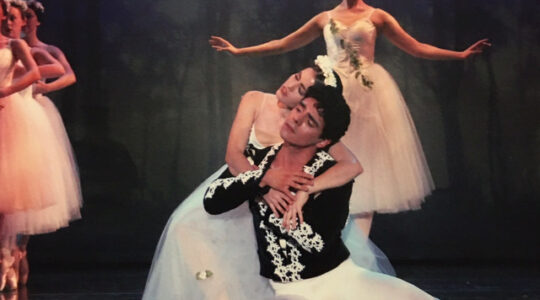 A group of ballet dancers perform on stage in a TCU Dance production. At the center, Domingo Estrada dressed in black and white kneels, holding a female dancer in his arms as she rests against him, wearing a white tutu.