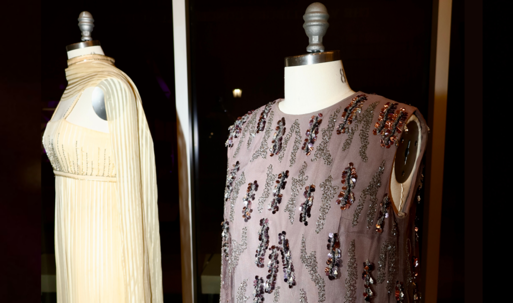 Photo of two dresses from the Historic Costume Collection. The dress in the foreground is purple with sequins, while the dress in the background is yellow with a matching silk scarf draped around the mannequin.